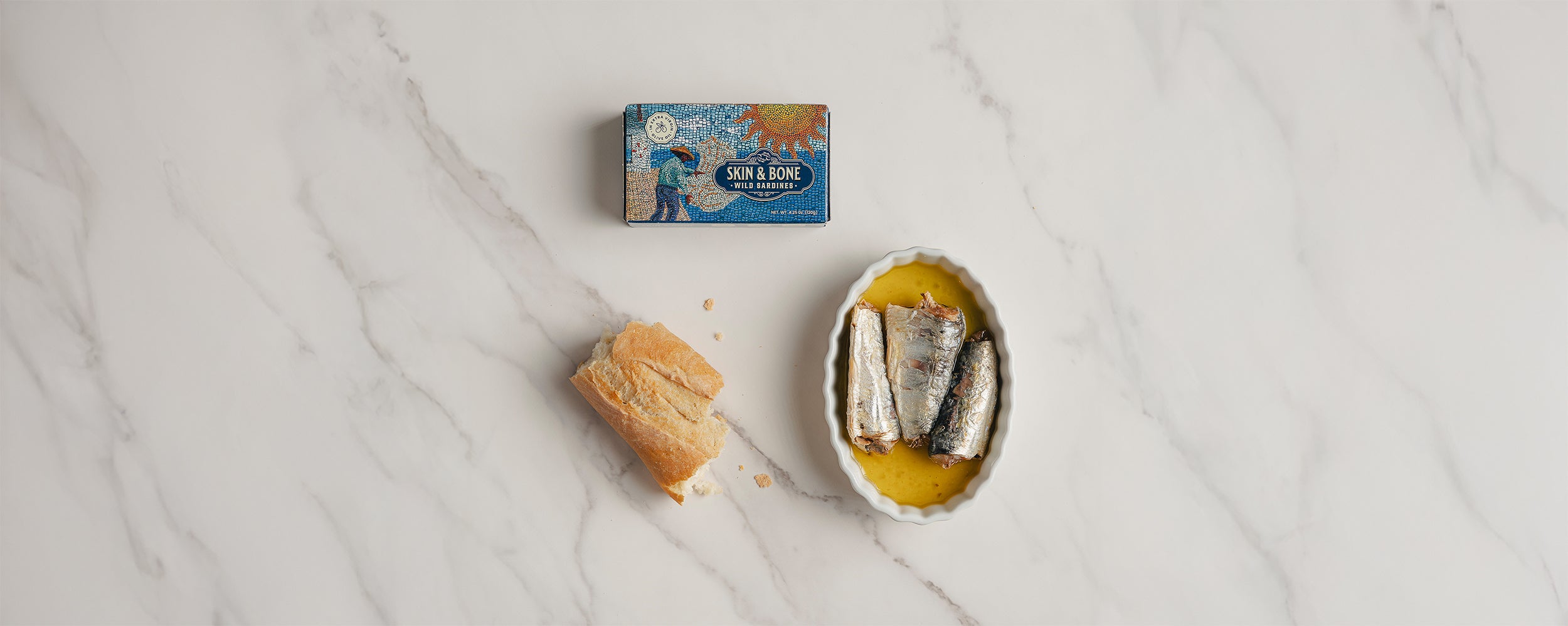Skin & Bone Wild Sardines tin next to a ramekin of sardines in golden olive oil and a piece of rustic bread on a white marble surface