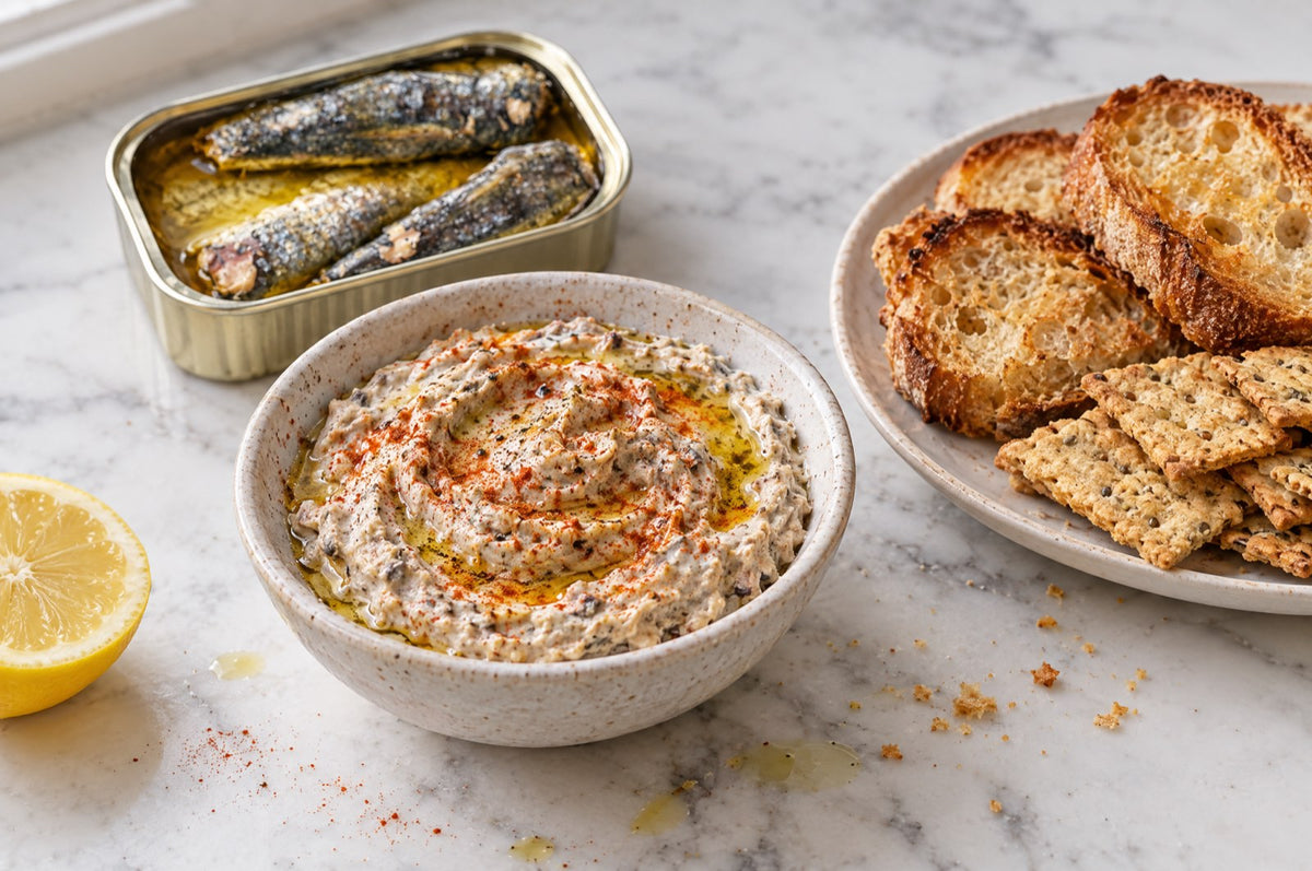 Bowl of whipped sardine dip with olive oil and smoked paprika, surrounded by crackers, toast and vegetables, next to an open tin of Skin & Bone sardines