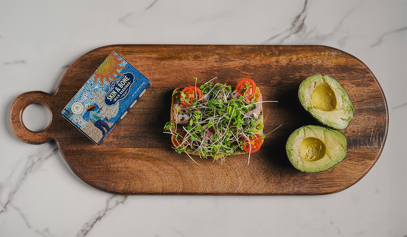 Skin & Bone Wild Sardines tin next to avocado toast with microgreens, tomatoes, and sliced avocado on a wooden serving board over marble countertop
