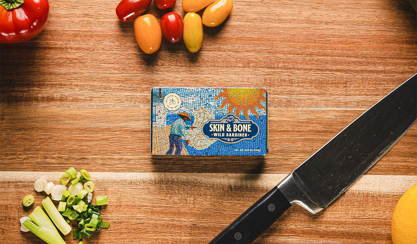 Skin & Bone Wild Sardines tin on a wooden cutting board surrounded by fresh vegetables, chef’s knife, and ingredients for meal prep